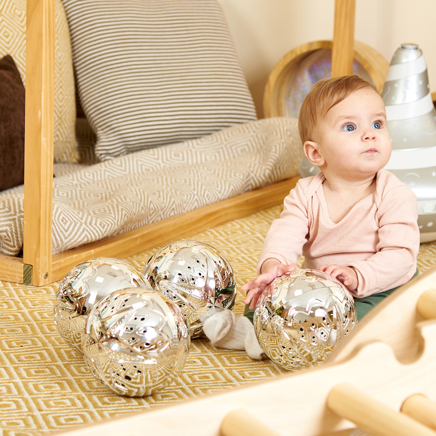 Bolas Sensoriais Prateadas Bebé sentado no tapete com bolas prateadas e almofada às riscas numa sala com estrutura de madeira