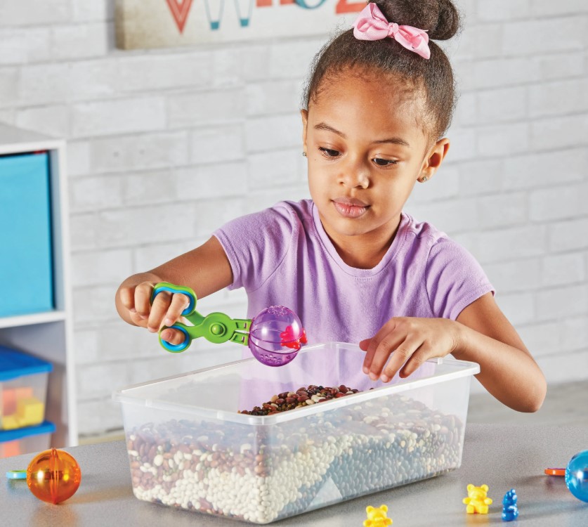 Tesoura bola - Handy Scoopers Criança com brinquedos e caixa plástica cheia de bolinhas coloridas numa mesa