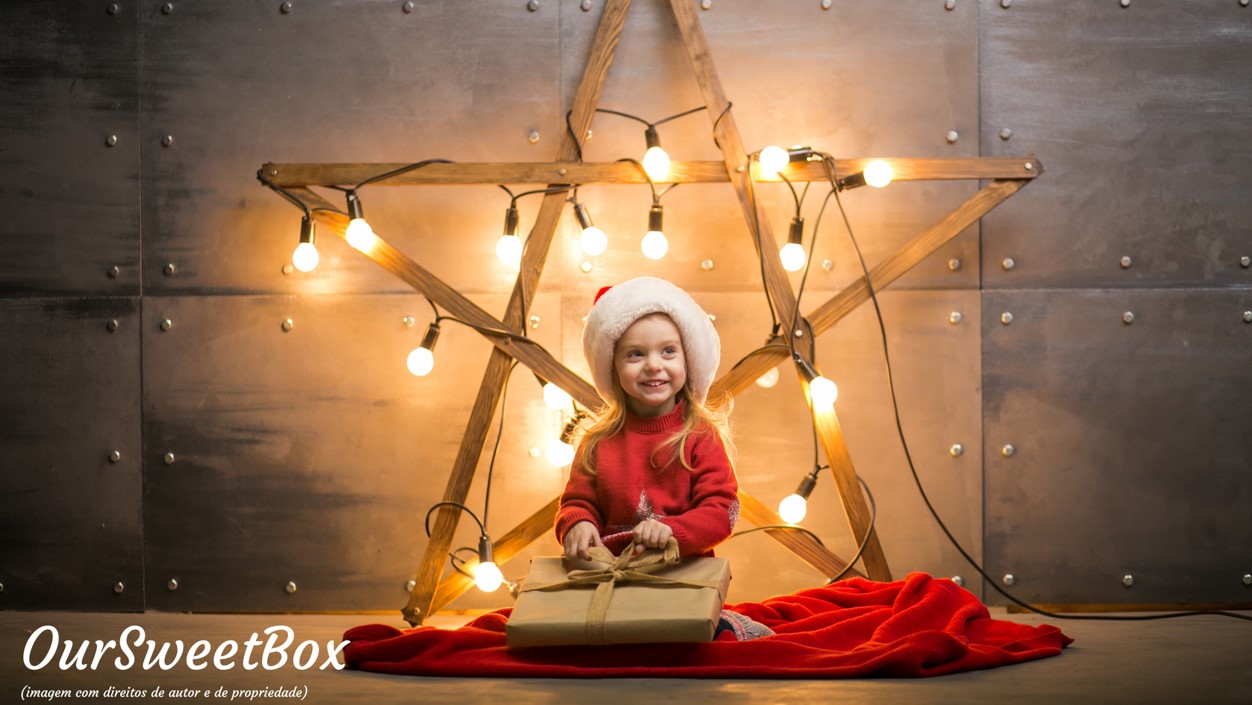 Criança sentada com gorro branco e camisola vermelha, estrela de madeira com luzes ao fundo