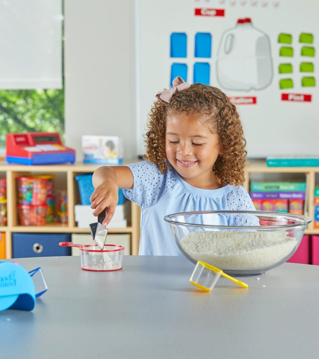 Chávenas de medição - Dois modelos Menina com vestido azul a mexer ingredientes numa mesa com tigela transparente e copo medidor numa sala de aula colorida.