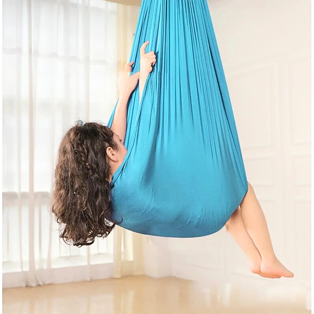 Rede Terapêutica Tipo Casulo - Therapeutic Hammock – Cocoon Criança dentro de saco suspenso azul em ambiente interior com chão de madeira e janelas grandes