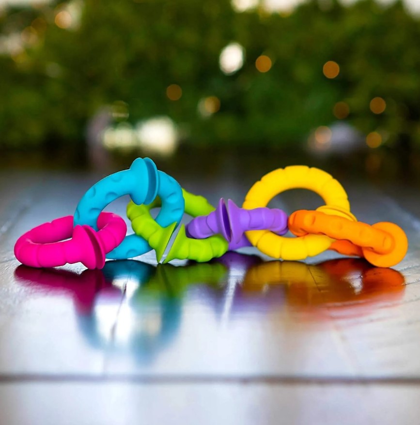 Anéis Sensoriais Texturados – Conjunto de 6 Brinquedos de anéis coloridos entrelaçados sobre piso de madeira com fundo verde desfocado