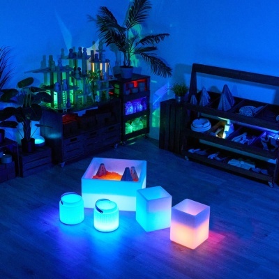 Sala com mobiliário escuro, plantas verdes e lâmpadas coloridas em forma de cubo e cilindro