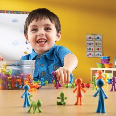 Menino a brincar com figuras coloridas de plástico sobre mesa com recipiente e parede amarela ao fundo.
