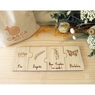 Quebra-cabeças educativo de madeira mostrando o ciclo da borboleta com texto explicativo, acompanhado de bolsa de pano natural com logotipo CASCA VIVA e plantas secas.