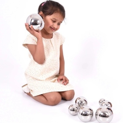 Menina ajoelhada com vestido branco segurando bola metálica prateada, rodeada por bolas metálicas