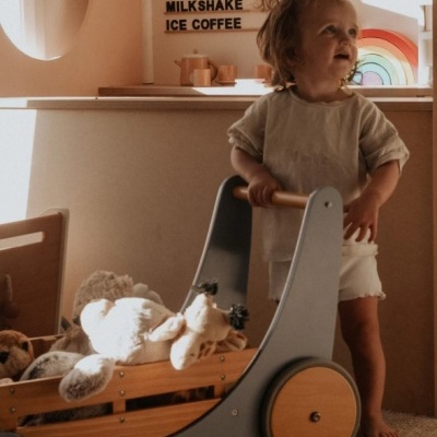 Criança com carrinho de brinquedo cheio de peluches e painel com textos e arco-íris ao fundo