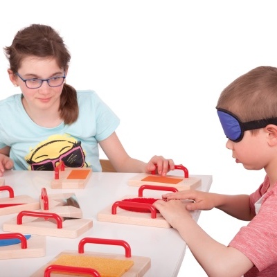 Crianças a explorar tábuas táteis com alças coloridas numa mesa branca