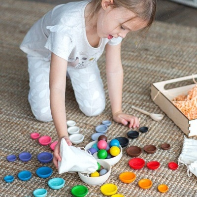Menina brincando com peças educativas coloridas num tapete com caixa de madeira ao lado.