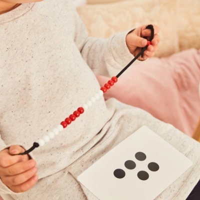 Criança segurando fio com contas vermelhas e brancas, com cartão branco com cinco pontos pretos na mão