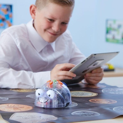 Robô educativo transparente com olhos e botões coloridos em tapete, menino com tablet ao fundo