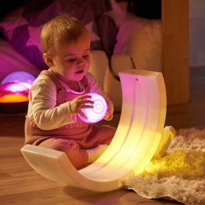 Bebé com bola luminosa rosa sentado ao lado de balanço curvado branco iluminado amarelo.