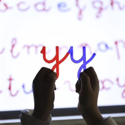 Mãos segurando letras 'y' vermelha e azul frente a quadro branco com letras do alfabeto coloridas.