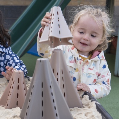 Criança a brincar com brinquedos triangulares em caixa de areia num parque infantil.