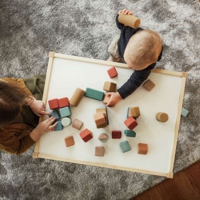 Duas crianças a brincar com blocos de madeira coloridos numa base branca com moldura de madeira sobre tapete cinza