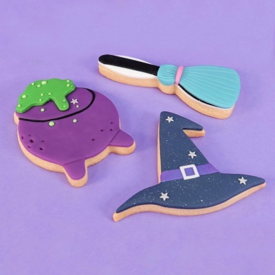 Bolachas decoradas com temas de bruxaria: caldeirão, vassoura e chapéu de bruxa, sobre fundo roxo.
