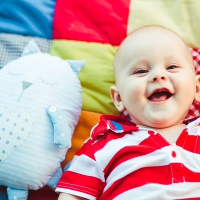 Bebé sorridente de polo às riscas ao lado de almofada coruja azul numa manta colorida