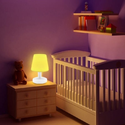 Quarto de bebé com berço branco, mesa de cabeceira com lâmpada acesa e urso de peluche, prateleiras com livros e brinquedos