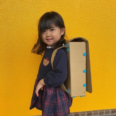 Criança com mochila de cartão castanho e botões azuis, roupa azul escuro e saia xadrez, contra parede amarela