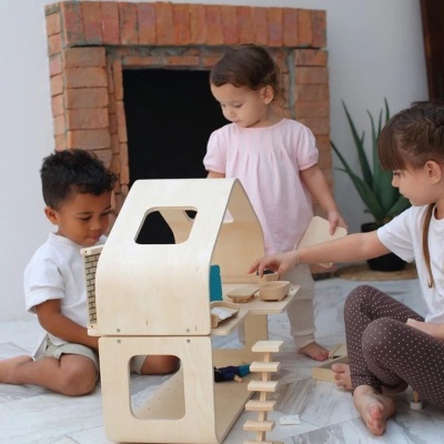 Crianças a brincar com casinha de madeira clara num chão interior, com lareira de tijolo ao fundo