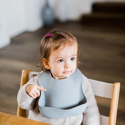 Bebé sentado com babete de silicone cinzento claro à mesa de madeira