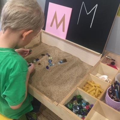 Criança a brincar com pedras e caixa de areia em mesa de madeira com vários materiais e quadro negro com letras M.