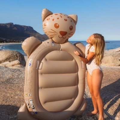 Colchonete insuflável castanho claro em forma de gato com manchas e bigodes perto do mar, criança loira ao lado