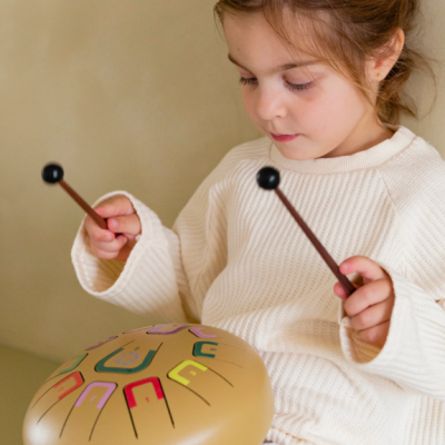 Criança com camisola branca a tocar instrumento musical tambor de língua bege com línguas coloridas.