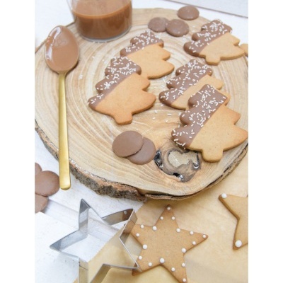 Biscoitos decorados em formas de árvore e estrela sobre disco de madeira e papel, com colher dourada e cortador de biscoitos