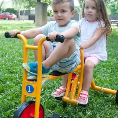 Crianças num triciclo amarelo num parque com relva e árvores