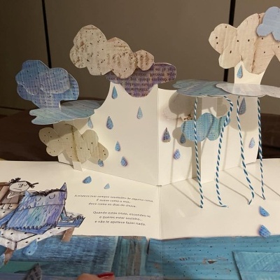 Livro pop-up com nuvens, gotas de chuva e peixe de papel em tons azuis e bege