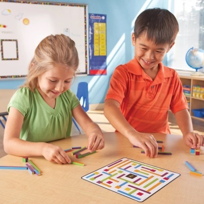 Duas crianças a jogar com peças coloridas e tabuleiro labiríntico numa sala de aula