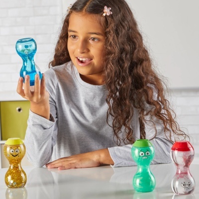 Menina segurando brinquedo azul em forma de ampulheta, mesa com brinquedos coloridos, parede branca de tijolos