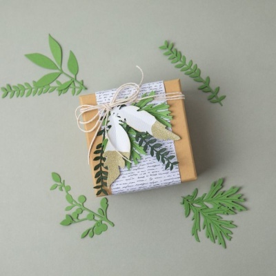 Caixa de presente decorada com elementos recortados em papel e laço em fio natural sobre fundo cinza com folhas verdes recortadas