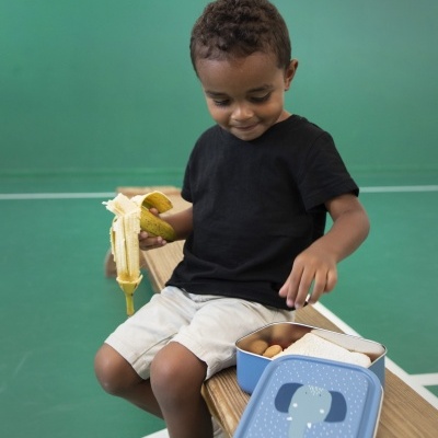 Menino com banana descascada e lancheira azul com elefante sentado num banco num campo desportivo verde