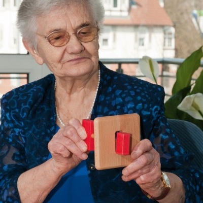 Senhora idosa segura brinquedo de madeira com peças vermelhas em interior com janelas e plantas