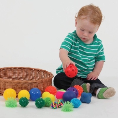 Criança brinca com bolas sensoriais coloridas junto a um cesto de verga em fundo branco