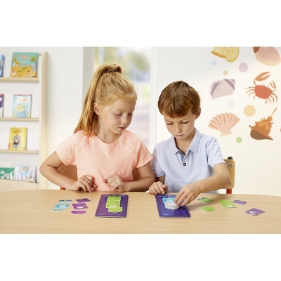 Crianças a jogar jogo de cartas com tabuleiros roxos e cartas coloridas numa mesa de madeira clara.