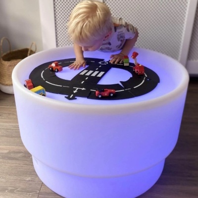 Mesa redonda iluminada LED azul com tapete pista e carros brinquedo onde menino loiro brinca