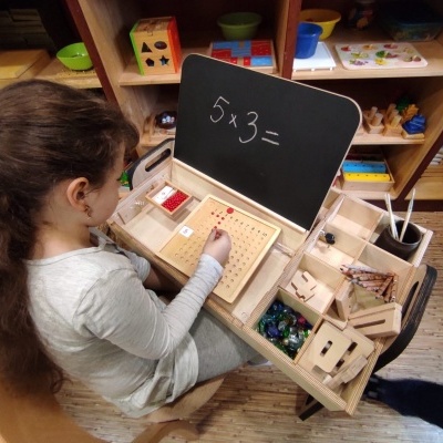 Criança com caixa educacional de madeira e quadro preto com equação 5x3=