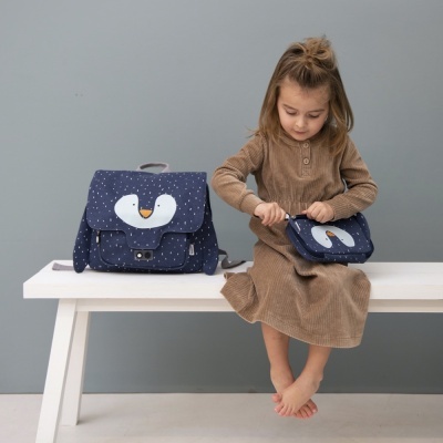 Mochila e carteira azul escuro com padrão de pontos e pinguim, menina com vestido castanho sentada numa bancada branca.