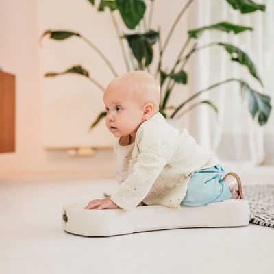 Bebé de roupa clara ajoelhado sobre almofada branca em interior com planta e móvel de madeira
