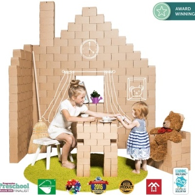 Crianças a brincar numa casa feita de blocos de cartão castanho com móveis e urso de peluche