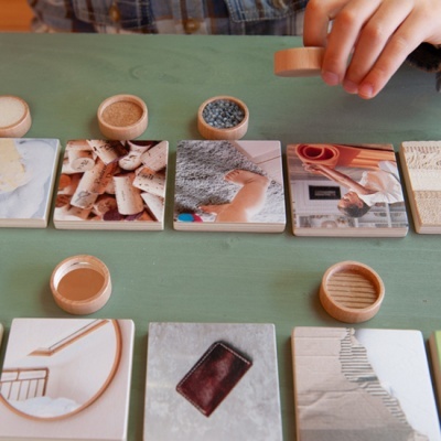Peças quadradas de madeira com imagens e recipientes redondos sobre mesa verde.
