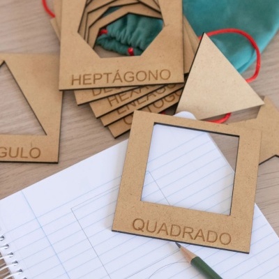 Molduras de cartão com formas geométricas e nomes em português sobre mesa de madeira com caderno aberto e lápis verde