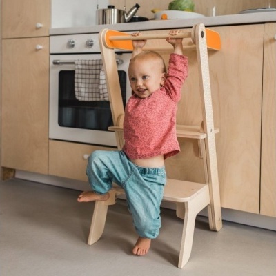 Bebé num escadinha de cozinha de madeira clara com proteções laranja, numa cozinha moderna