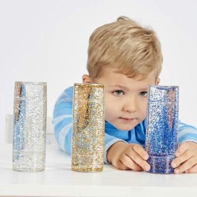 Menino observando três garrafas transparentes com glitter nas cores branco, dourado e azul sobre mesa branca