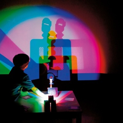 Criança a projetar sombras coloridas numa parede com objetos e luzes coloridas