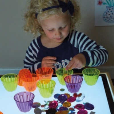 Menina loira a brincar com copos de plástico coloridos e pedras numa mesa luminosa