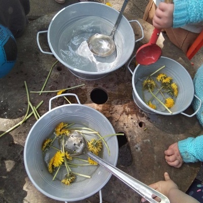 Três baldes metálicos com água e flores amarelas, conchas metálicas e uma criança com colher vermelha